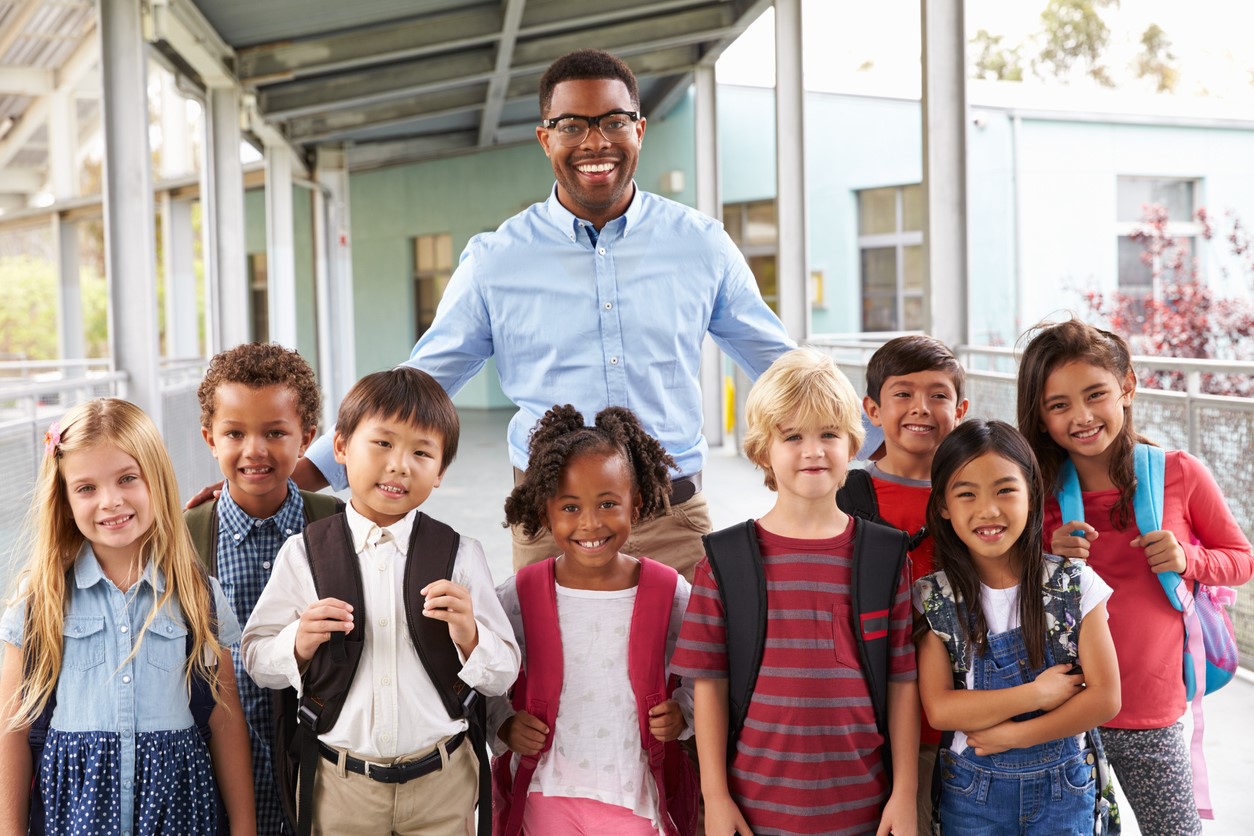 african american male teacher with eight young students from diverse backgrounds standing outside school