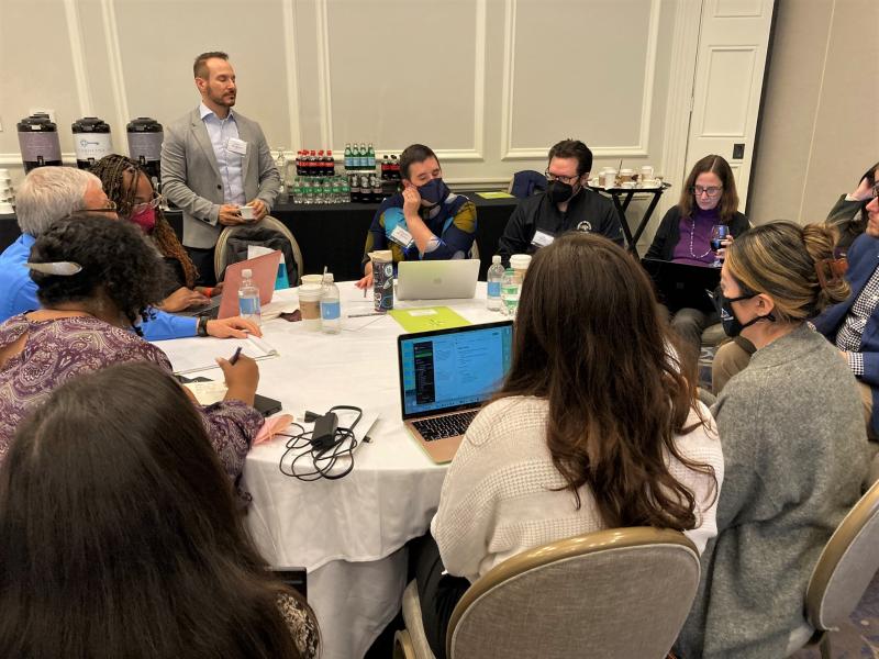 data camp attendees sit and stand around large round table, some wear face masks