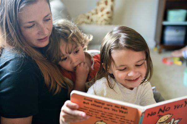 caucasion mom holding small child in lap while watching another small child read a picture book