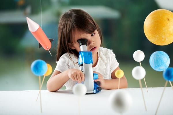Pre-school aged girl looking through microscope and enjoying scientific activity for preschoolers, blurred background