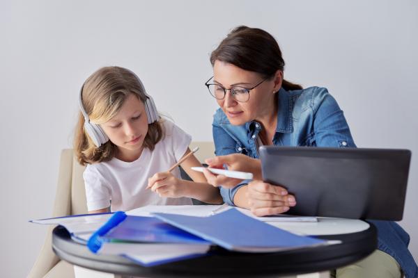 mom helping daughter using digital tablet; daughter at left in headphones, writes in notebook with pencil