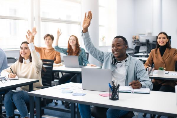 A group of students sits in a classroom listening to their professor off-screen. They are taking notes. One man of African descent raises his hand to ask a question.