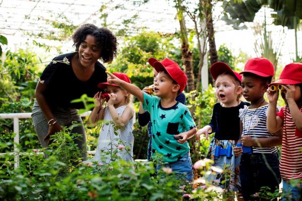 teacher and six young students wearing red hats inside a greenhouse looking at plants