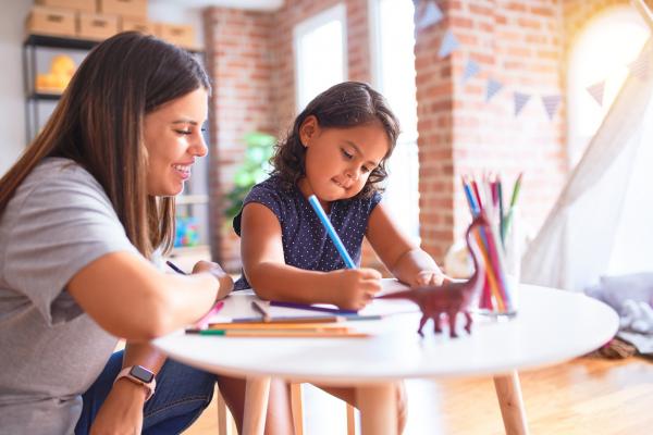 female teacher watching and helping toddler girl using colored pencils at kindergarten