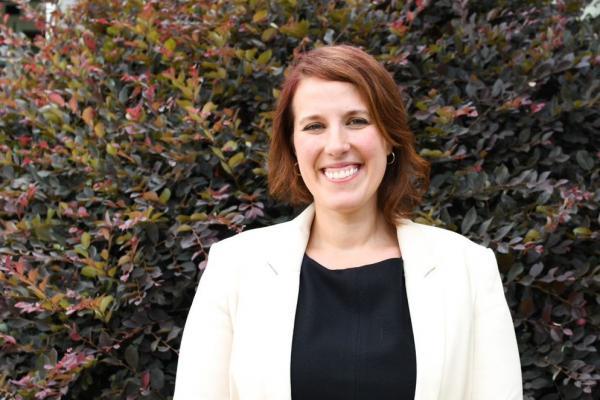 Julie Austen; woman with shoulder-length hair wearing black top and white jacket stands in front of large shrub and is smiling widely into camera