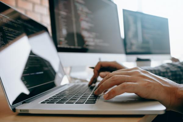 someone's hands type on a laptop's keyboard with two monitors in the background