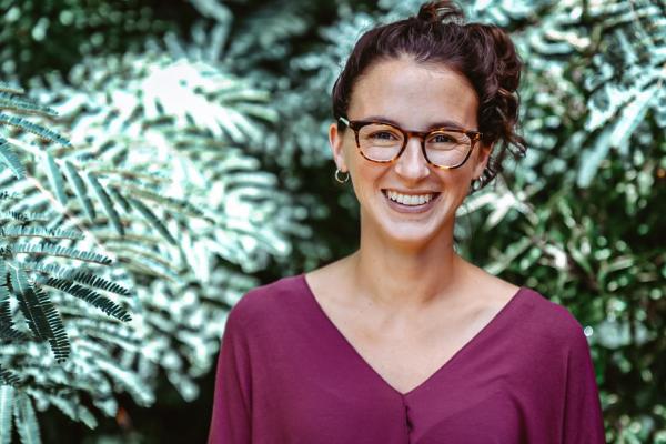 Kelsey Thompson headshot; woman in purple v-neck top with dark hair and glasses stands outside in front of hedge