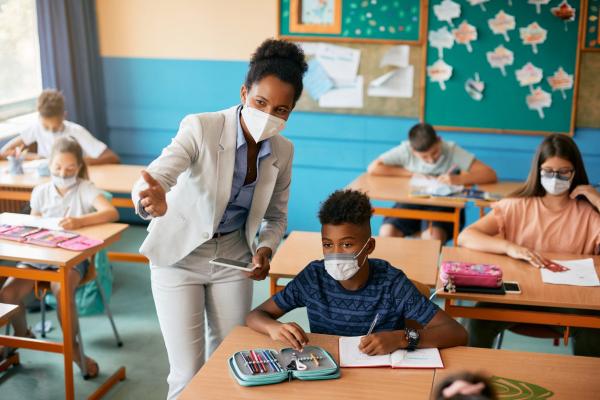 teacher wearing face mask talking with students wearing facemasks who are seated at desks in a small middle school classroom