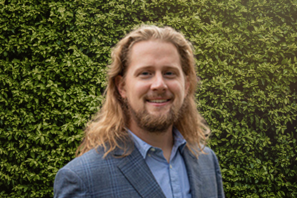 Nathan Jorgensen; man with long blonde/brown hair stands in front of green hedge