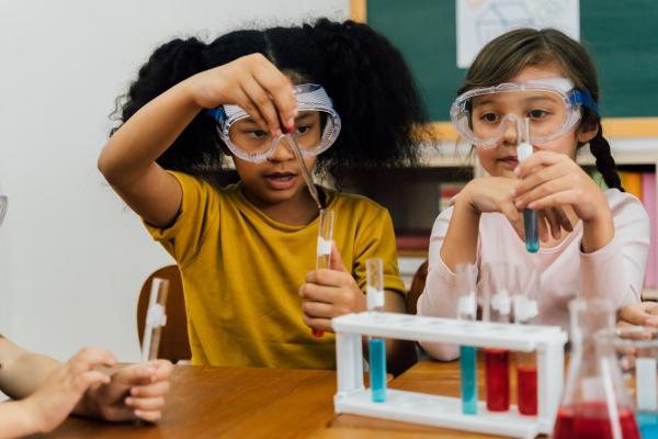 girls in safety goggles sitting at table mixing liquids in vials during chemistry lesson at primary school