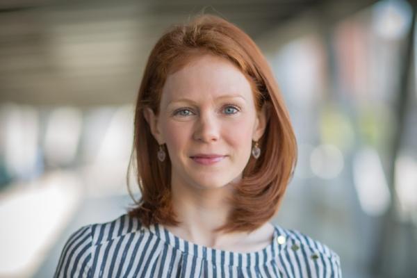 Samantha Schilling, MD, MSHP; woman with shoulder-length red hair wearing blue and white top smiles at camera in front of fuzzy building background