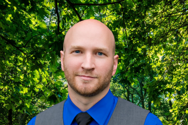 adam holland; man with shaved head and light-colored facial hair wearing a blue shirt and gray vest stands outside in front of lush greenery