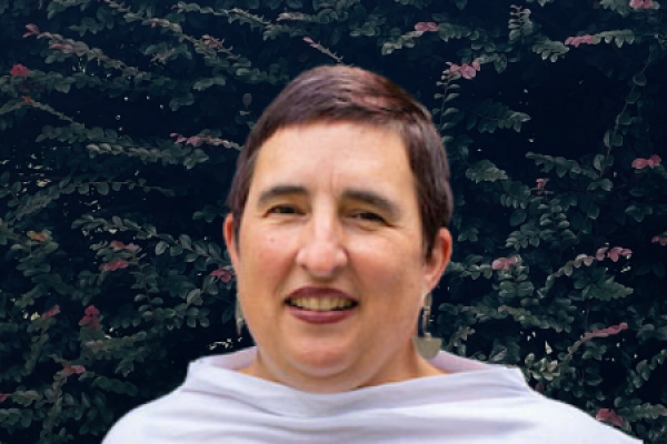 allison demarco headshot; woman with close-cropped dark hair wearing a white cowl-neck top stands in front of dark purple shrubbery