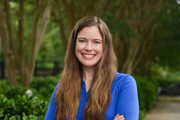 Ann Sam - woman with long brown hair wearing blue shirt stands outside in front of trees and shrubbery