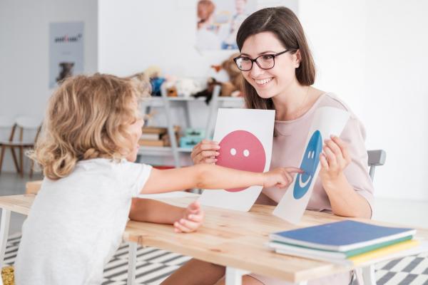 classroom setting with young student with curly blonde hair points to smiling blue face image on paper held by woman with long dark hair and glasses