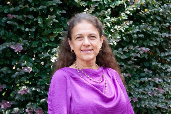 Ayse Belger; woman with long brown hair and wearing a raspberry blouse stands in front of a large green hedge