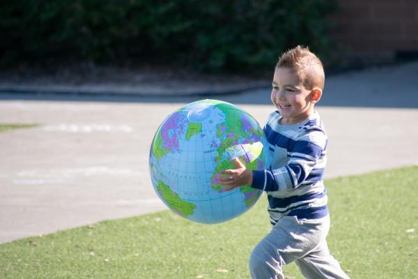 young boy with sandy brown hair and wearing a blue and white stripe top with gray pants carries an inflatable globe