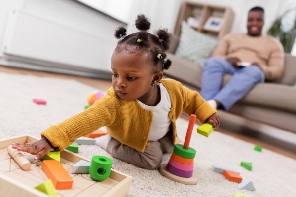 african american baby girl playing with toy blocks at home with father in background on sofa