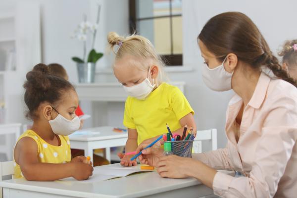 early child care classroom during COVID pandemic; two young students in masks interact with teacher wearing mask