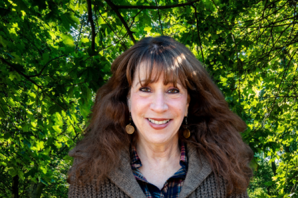 denni fishbein headshot; woman with long wavy hair standing in front of full, green trees smiles at camera