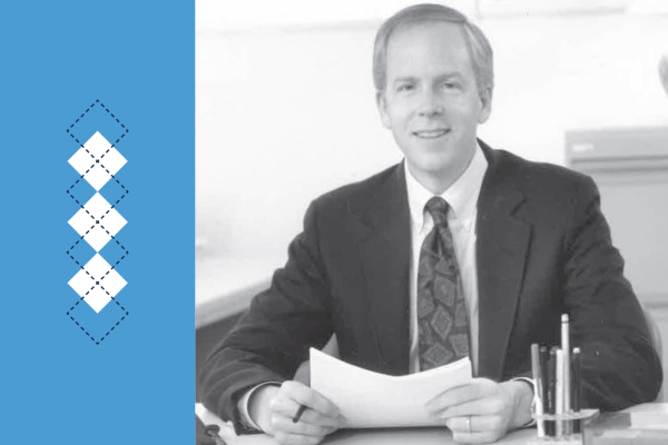 Black and white photo of Don Bailey sitting at a desk with Carolina Blue accent at left and argyle pattern