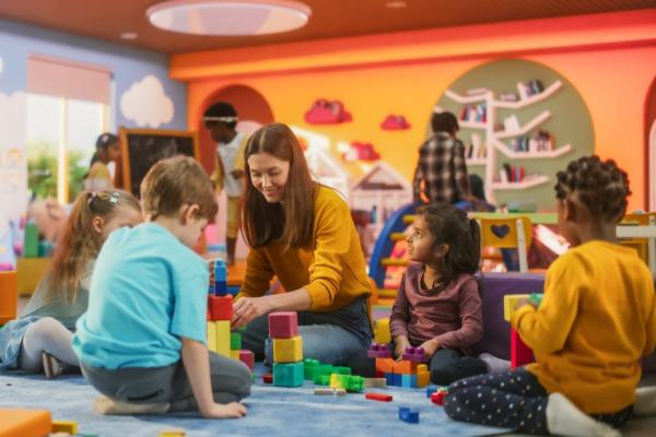 Cheerful diverse kids playing together with colorful building block toys with a female teacher in a modern daycare center