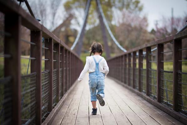 young girl with dark hair in pigtails is wearing white top and light blue overalls while running across a large bridge