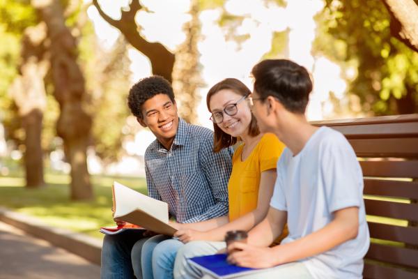 two male and one female grad students sitting on college campus bench with trees in background