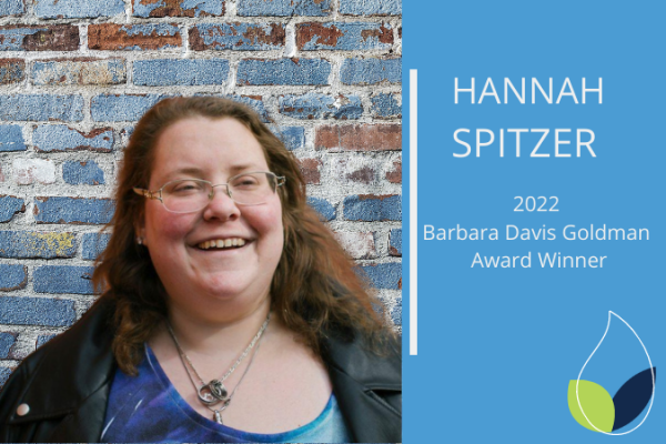 hanna spitzer smiles at camera; young woman with brown hair and glasses standing in front of wood panelled wall smiles at camera with  text  Hannah Spitzer 2022 Barbara Davis Goldman Award WInner