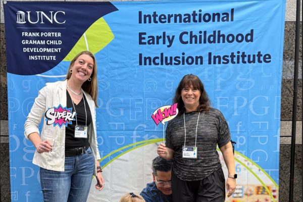 two women stand in front of the UNC FPG International Early Childhood Inclusion Institute sign during the 2025 conference