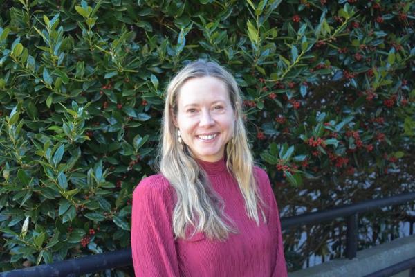 Jessica Amsbary; woman with long blond hair and wearing a magenta shirt stands in front of large shrub and smiles at camera