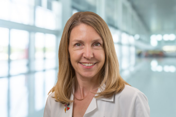 Kori Flower; woman with shoulder length blond hair wearing lab coat stands in bright, window-filled  hallway