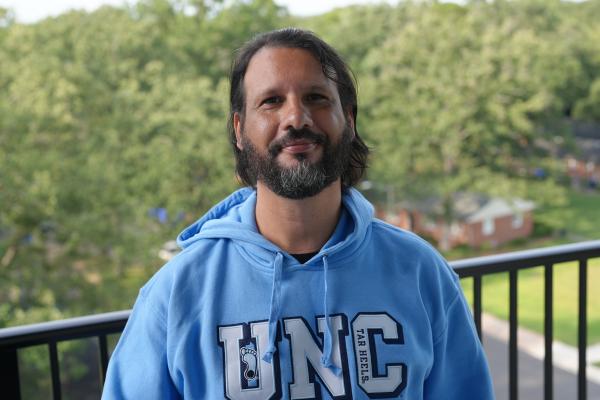Lucelmo Lacerda, a man with dark hair and beard, wearing a UNC carolina blue sweatshirt stands on outdoor balcony with trees in background
