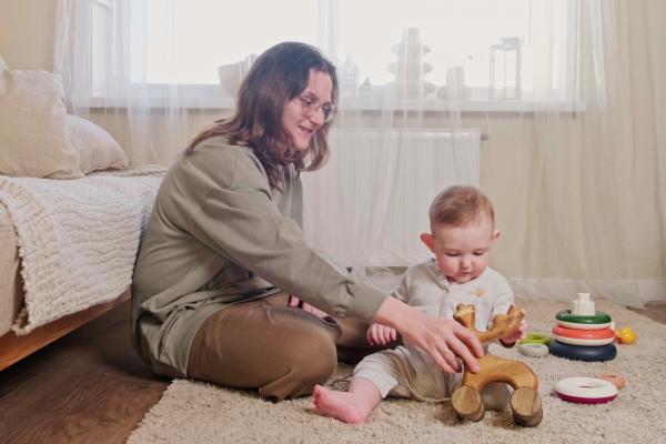 A toddler boy and mother play with a wooden moose toy on the carpet in their home. 