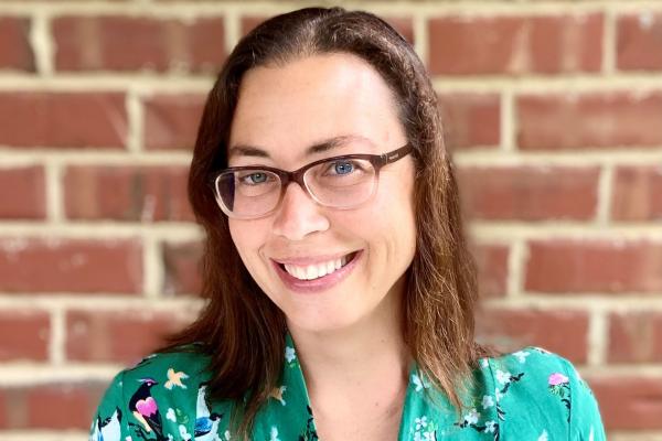 Nissa Towe-Goodman; woman with dark hair and glasses wearing green blouse stands in front of brick wall