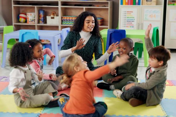 teacher interacting with children in pre-K classroom; sitting in a circle on colorful block mat with small tables and chairs in background