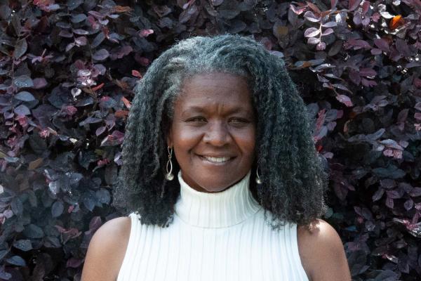 headshot of schatzi mccarthy; woman with shoulder-length hair wearing white turtleneck sweater standing in front of purple tree