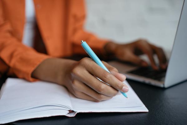 student wearing orange jacket sitting at desk writing on notepad beside laptop