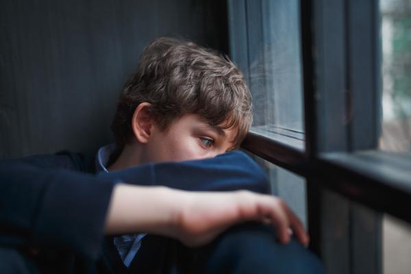 Pensive teenage boy with medium brown hair and blue eyes in a navy sweatshirt and jeans sits by a window partially covering his face with his arms and hands.