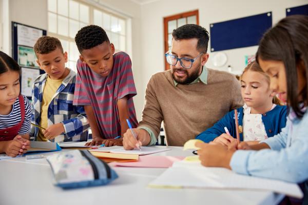 male teacher wearing eyeglasses sits at table surrounded by students and checks their work in notebooks