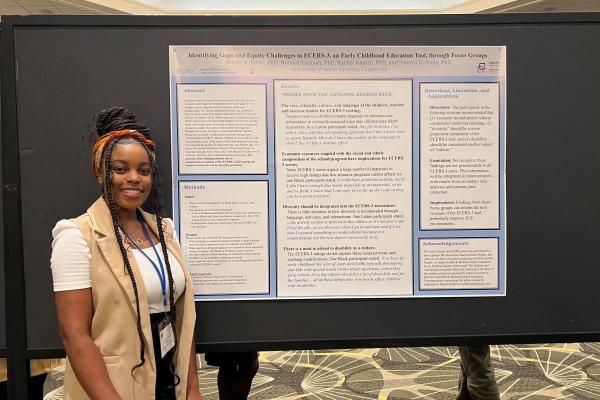 Nicole Telfer presents poster during Eastern Psychological Association Conference; woman with long braids wearing white blouse and tan vest stands beside large presentation poster