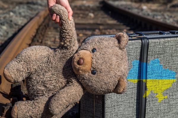 beige teddy bear held aloft above railroad tracks with suitcase and Ukraine country outline overlayed on top