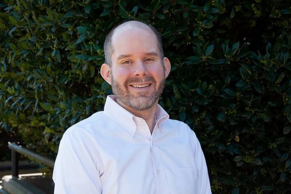 will aldridge; man with beard wearing white button-down shirt stands in front of shrubbery