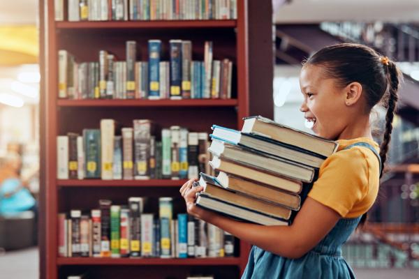 Young girl carries large stack of books through library