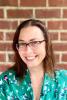 Nissa Towe-Goodman; shoulders up photo of woman with dark hair and glasses wearing a green print blouse standing in front of brick wall