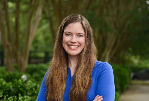 Ann Sam; woman with long chestnut hair and wearing a blue top stands outside smiling at camera