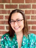 Nissa Towe-Goodman; shoulders up photo of woman with dark hair and glasses wearing a green print blouse standing in front of brick wall