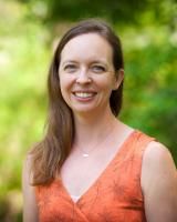 Stephanie Rszka, woman with auburn hair wearing orange top, stands outside in front of greenery